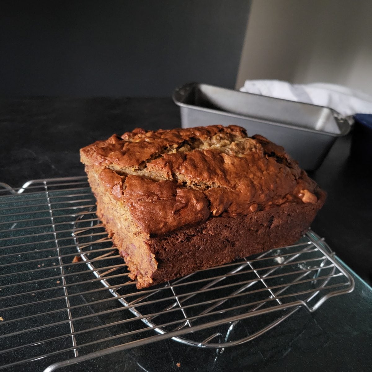 A loaf of banana bread cools on a metal rack, with an empty loaf pan and a white cloth in the background.