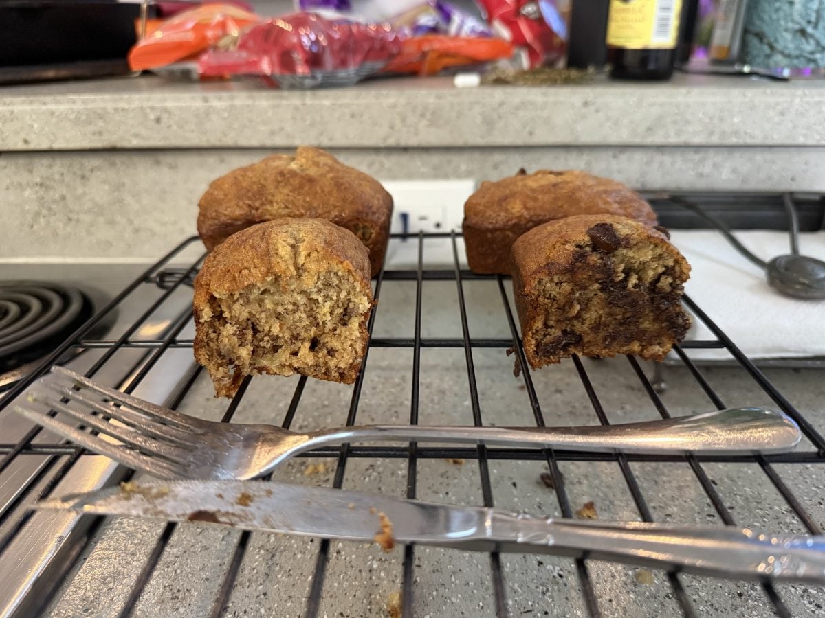 Four mini banana bread loaves, two sliced in half to show the inside, sit on a cooling rack with two forks in front; one loaf contains chocolate chips.