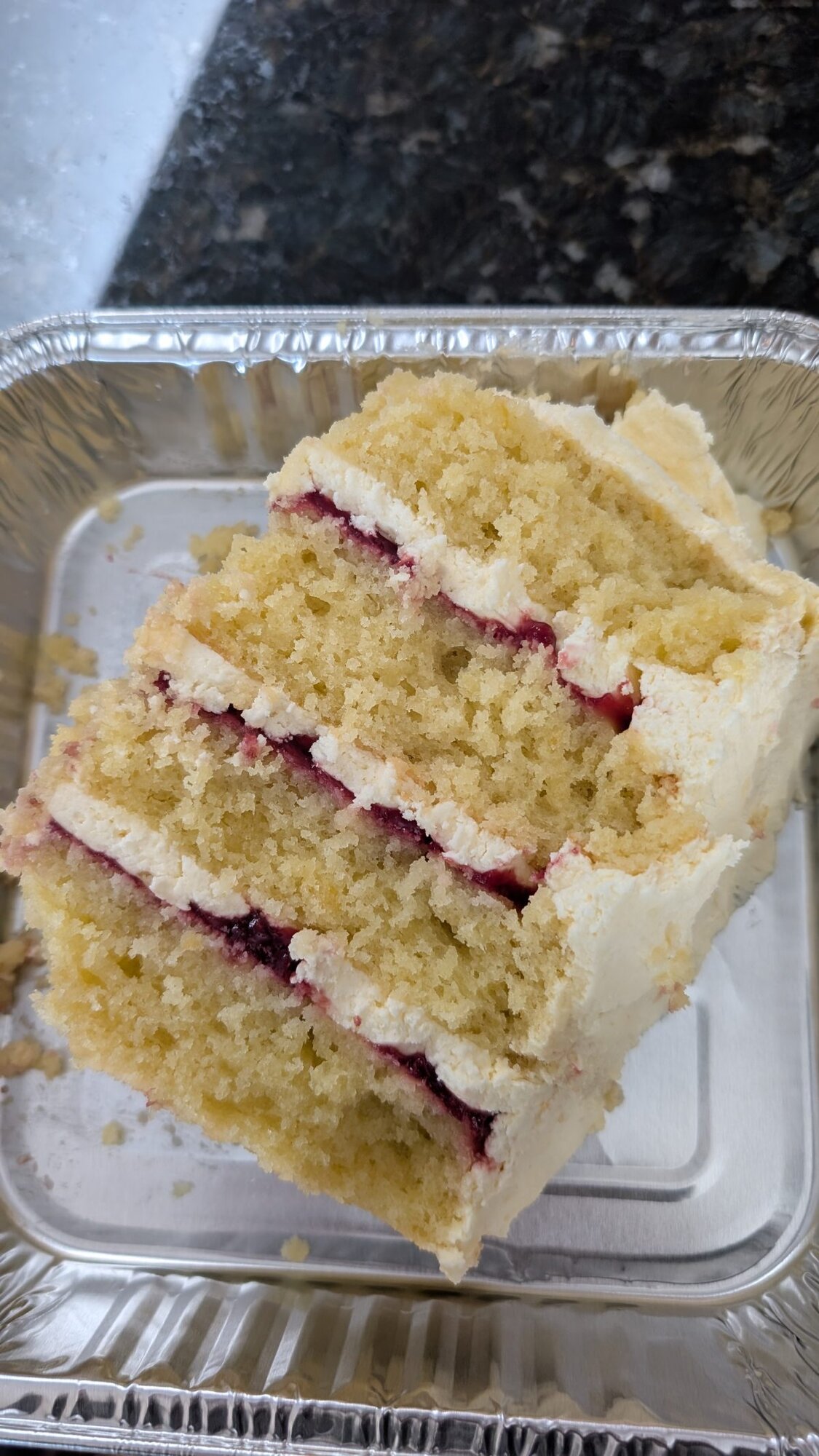 A slice of layered yellow cake with white frosting and a red jam filling in a rectangular aluminum container on a dark countertop.