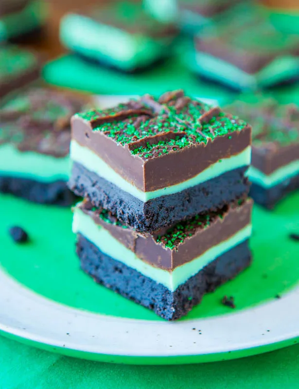 Two stacked chocolate mint brownies with three distinct layers and green sprinkles on top, placed on a white plate with more brownies in the background.