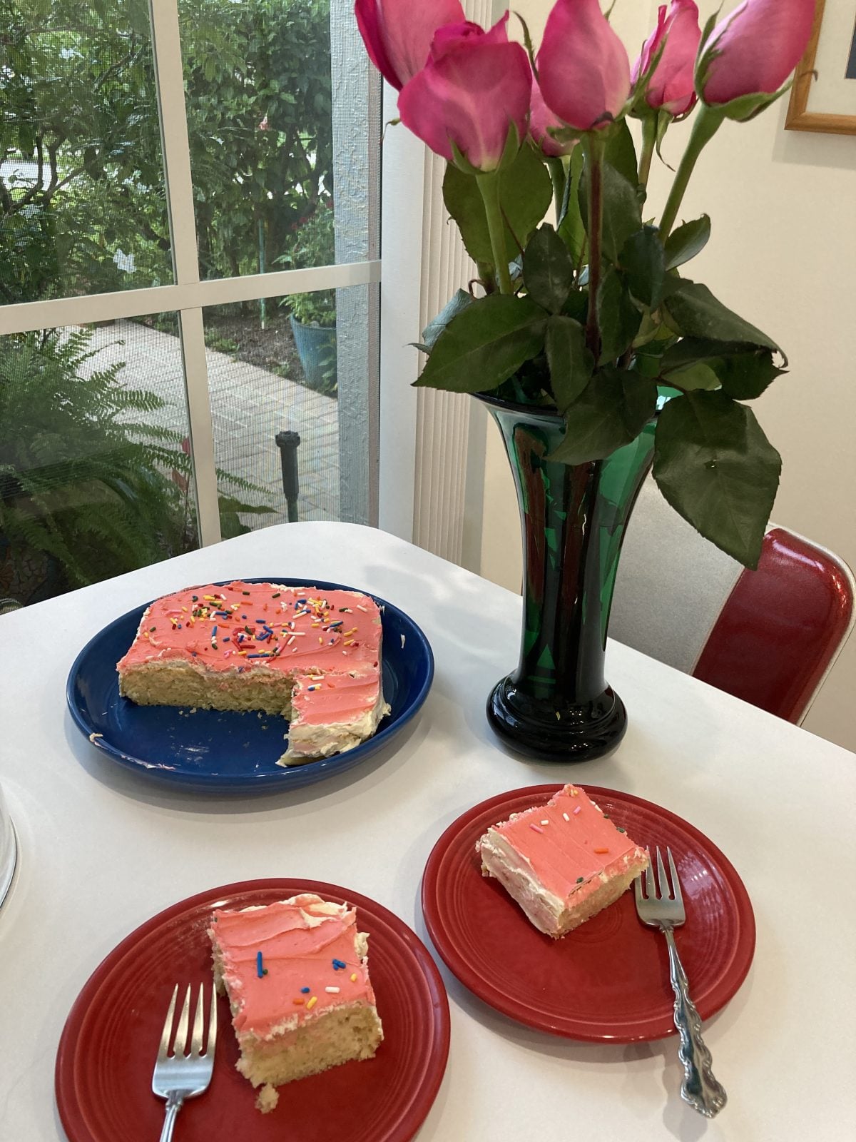 A table with a vase of pink roses, a partially eaten rectangular cake with pink frosting and sprinkles on a blue plate, and two slices of cake on red plates with forks.