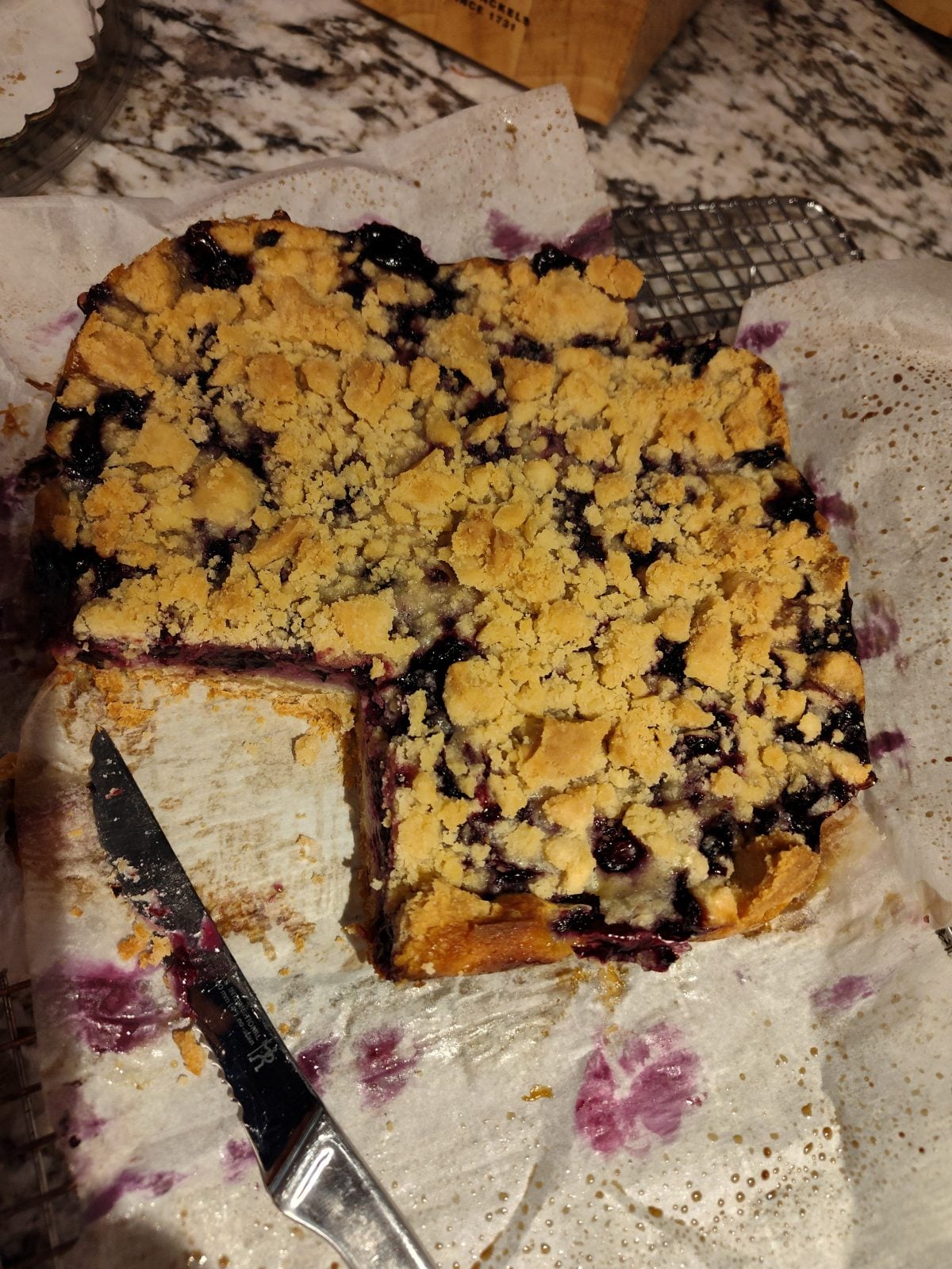 A square blueberry crumb cake with a slice removed, sitting on parchment paper. A knife rests nearby on a wire rack.