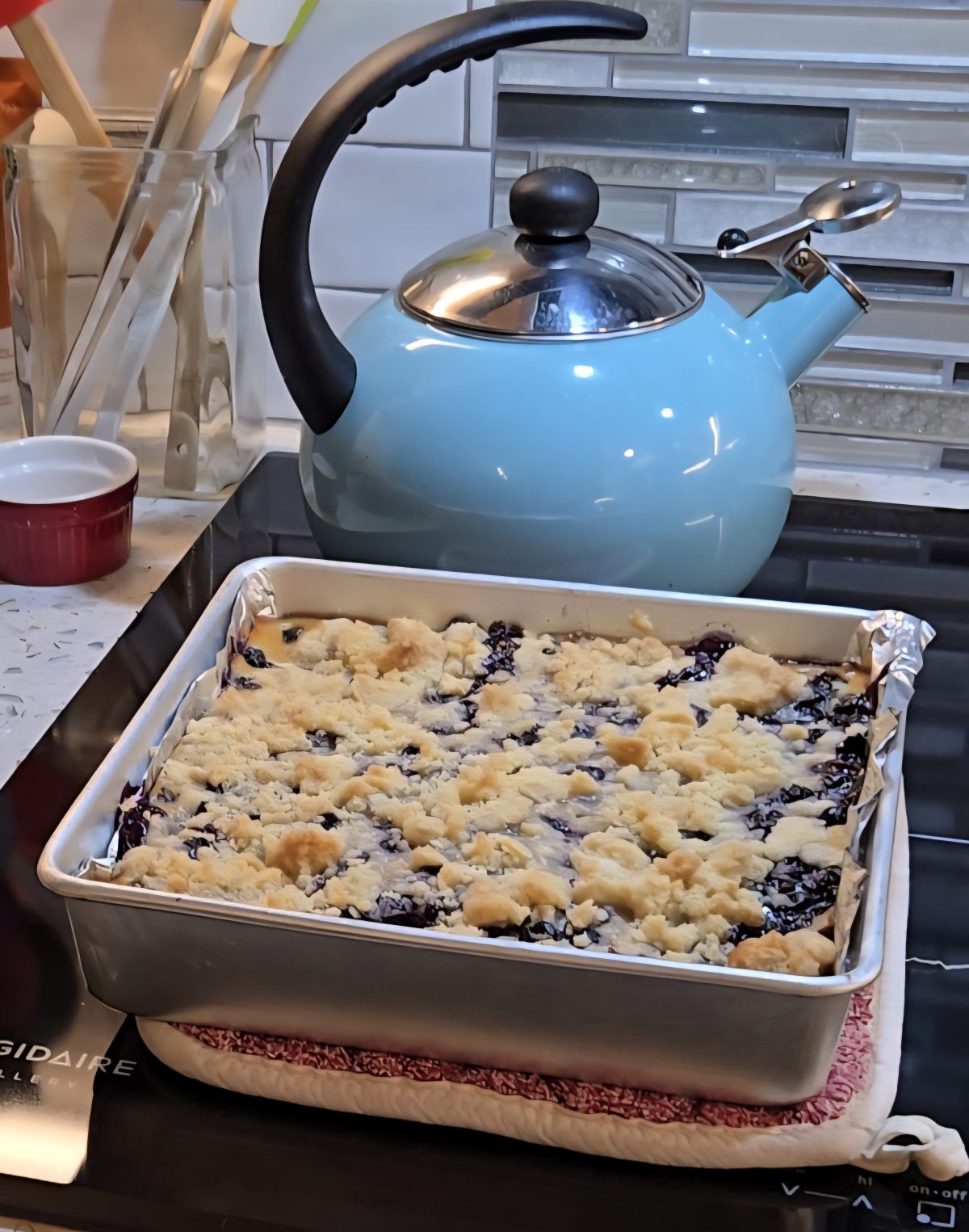 A square pan of blueberry crumb bars cools on a stovetop, with a blue kettle and kitchen utensils visible in the background.