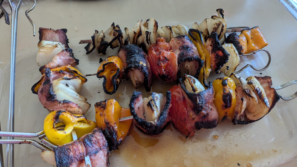 Skewers with grilled onions, yellow bell peppers, and pieces of meat, lightly charred and arranged on a glass tray.