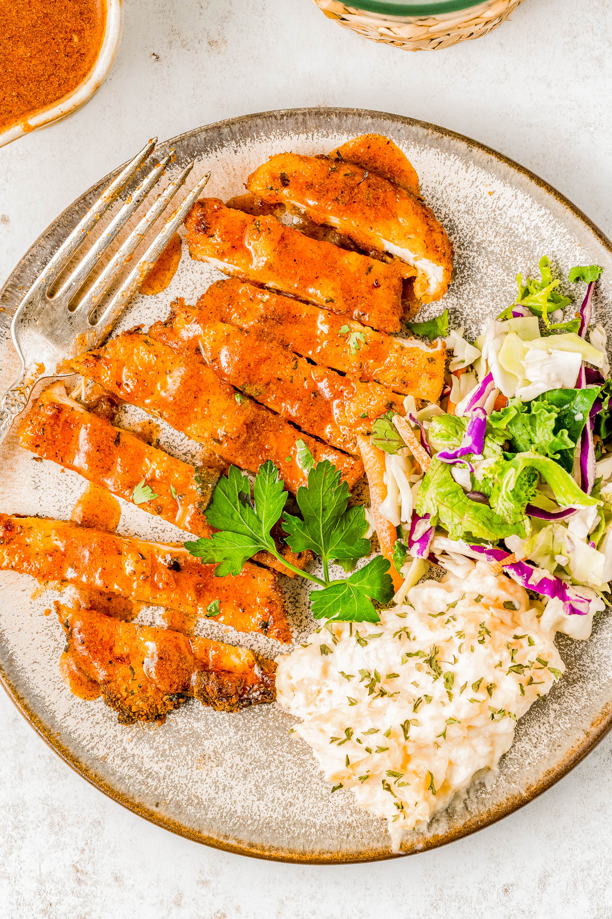 A plate with sliced grilled chicken topped with sauce, a side of coleslaw, and a mixed greens salad, garnished with parsley. 