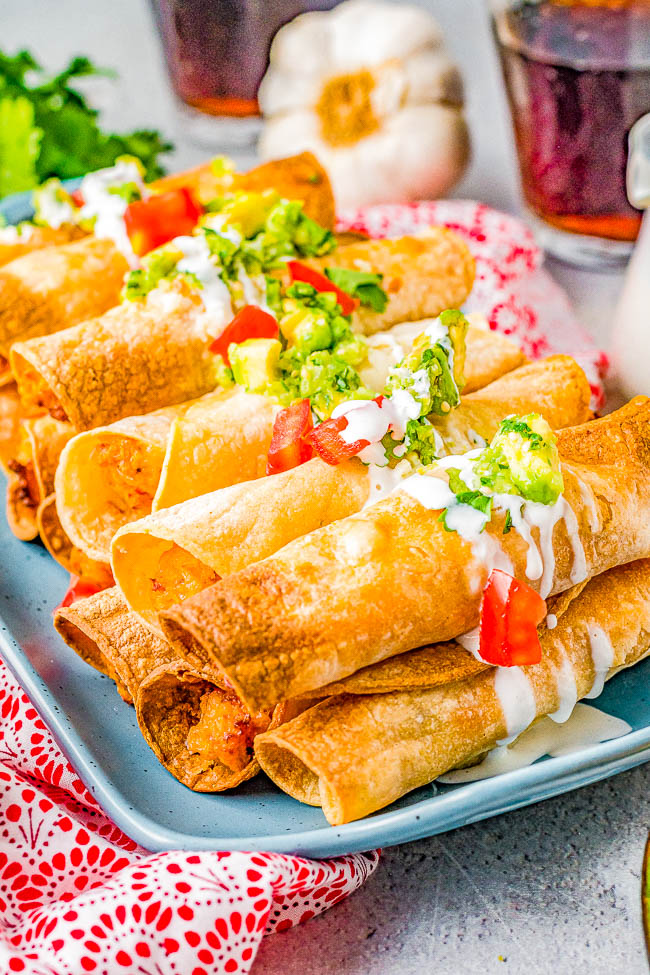 A plate of crispy taquitos topped with guacamole, diced tomatoes, and drizzled with white sauce.