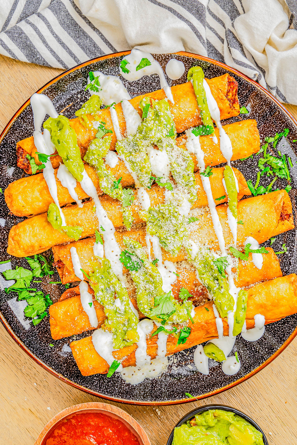 A plate of crispy taquitos topped with green sauce, white drizzle, and sprinkled cheese.