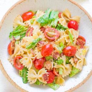 A bowl of bowtie pasta salad with cherry tomatoes, lettuce, bacon bits, and a light dressing, with additional ingredients scattered around the bowl.