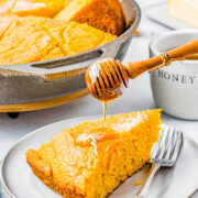 A slice of sweet potato cornbread on a plate with a fork on the side and honey being drizzled on top.