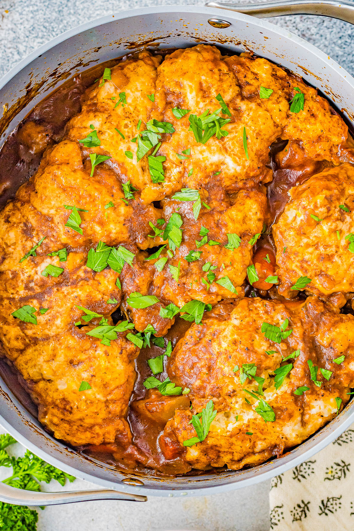 A pot of chicken and dumplings in a rich, brown sauce, topped with chopped parsley.