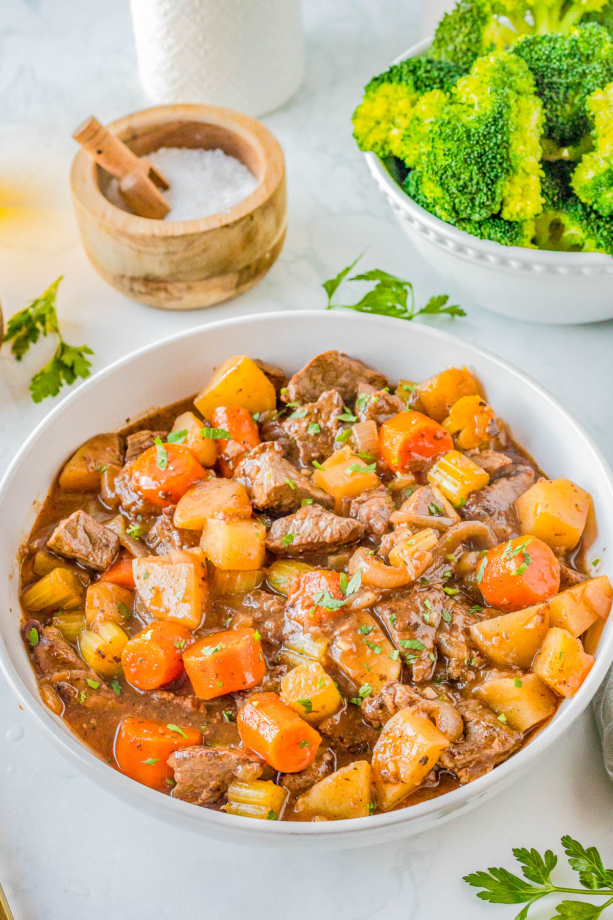 A bowl of beef stew with chunks of beef, carrots, potatoes, and celery. A side of cooked broccoli and a salt container are in the background.