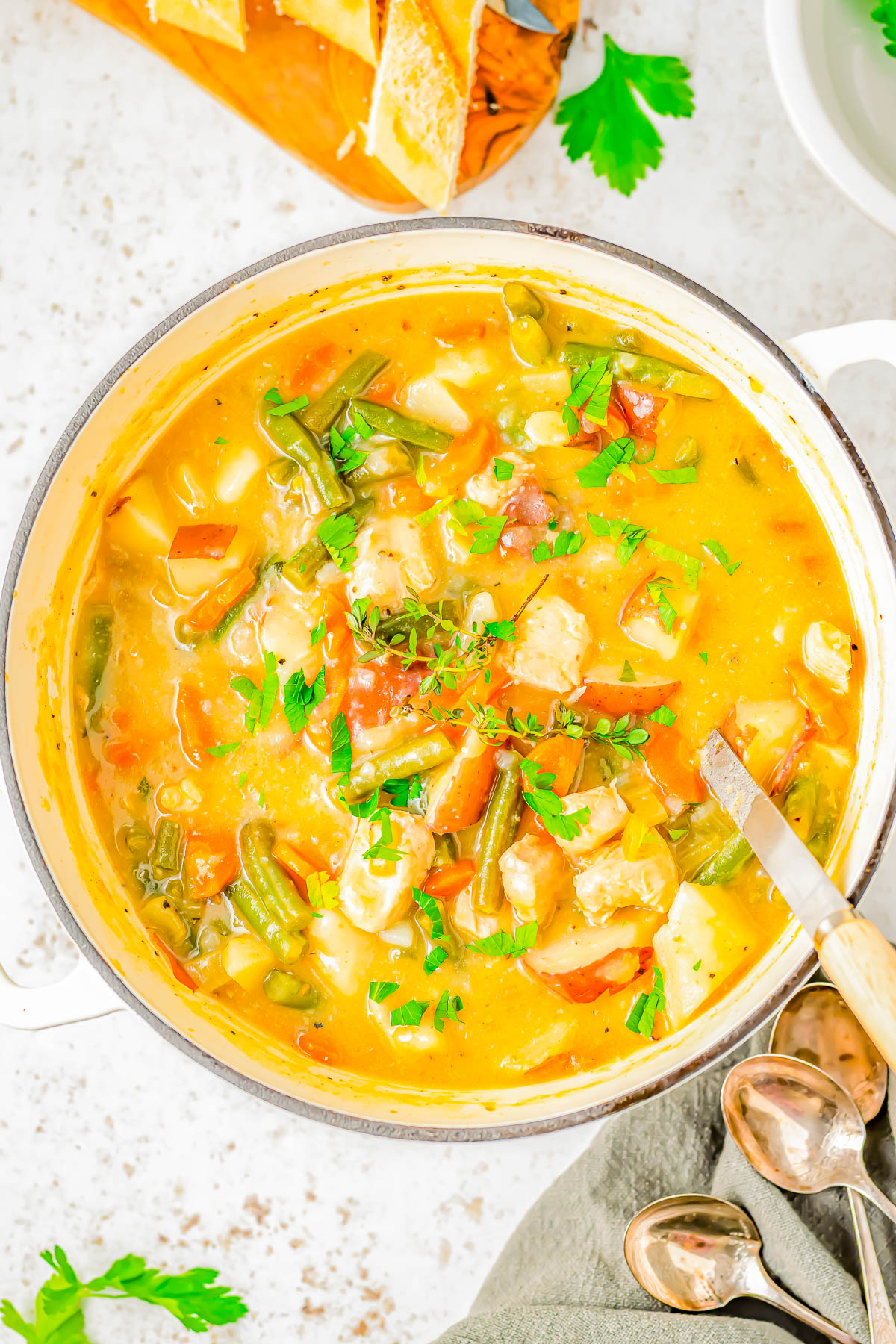 A pot of vegetable soup with green beans, tomatoes, and herbs. Sliced bread and parsley are nearby.