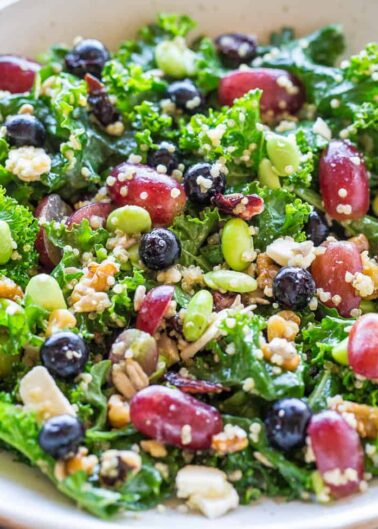 A colorful kale salad with red beans, blueberries, peas, grains, and crumbled cheese in a white bowl.