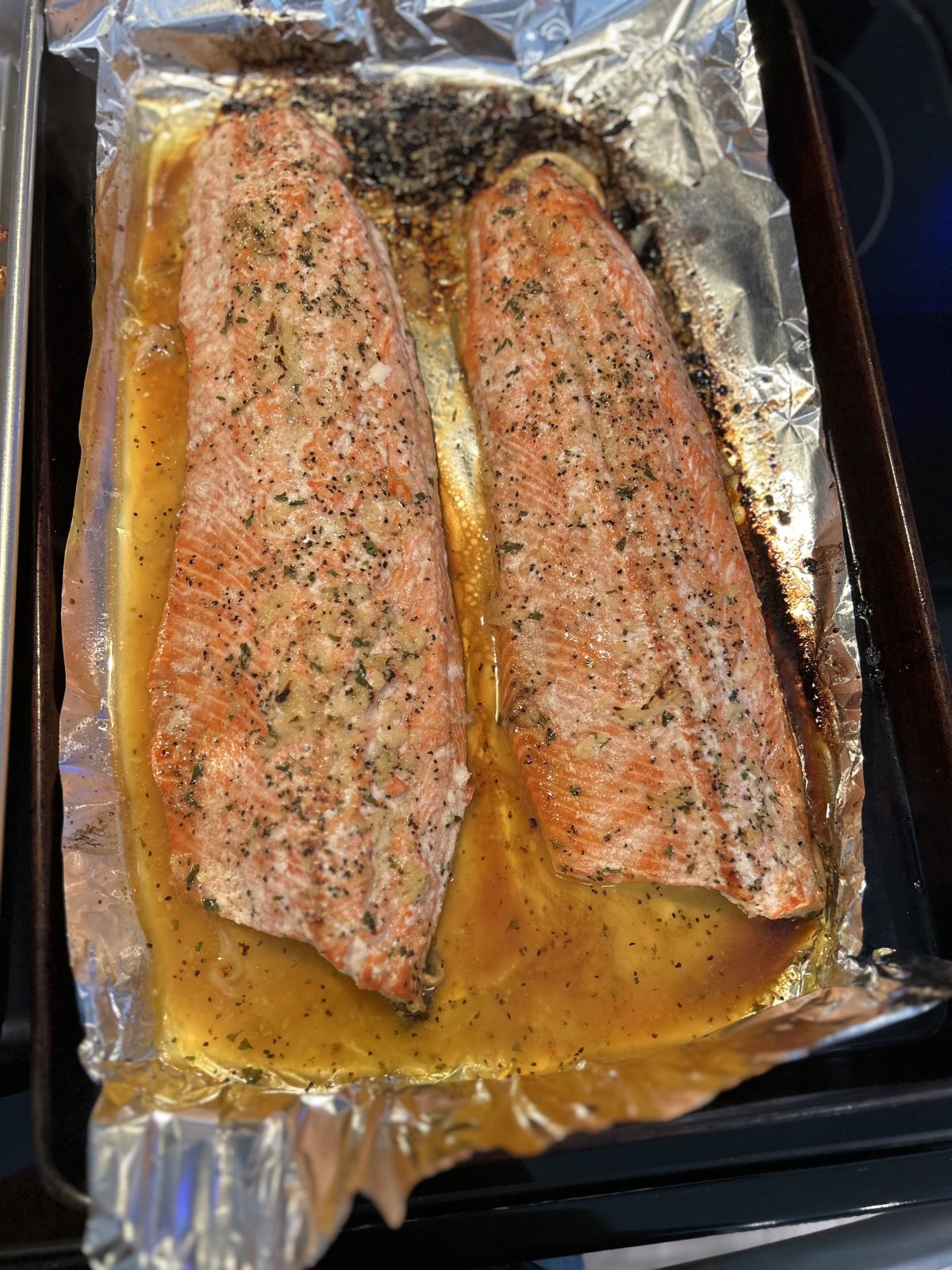 Two seasoned salmon fillets baked on a foil-lined tray, with visible juices and herbs surrounding the fish.