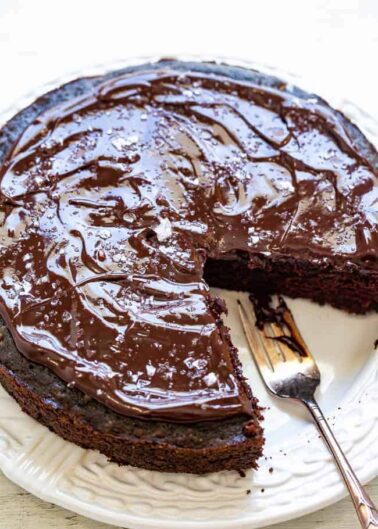 A chocolate cake with a glossy chocolate glaze, with one slice partially removed, placed on a white plate with a fork.