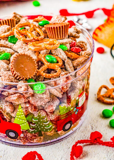 A festive container filled with a mix of holiday-themed candies and snacks, including chocolate pieces, pretzels, and coated cereals.