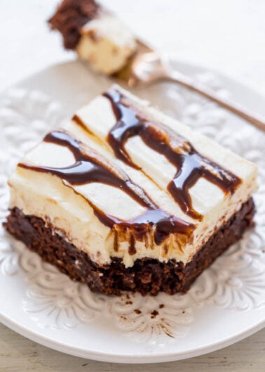 A slice of layered dessert with a brownie base, topped with cream cheese layer and drizzled with chocolate and caramel sauce on a white ornate plate.