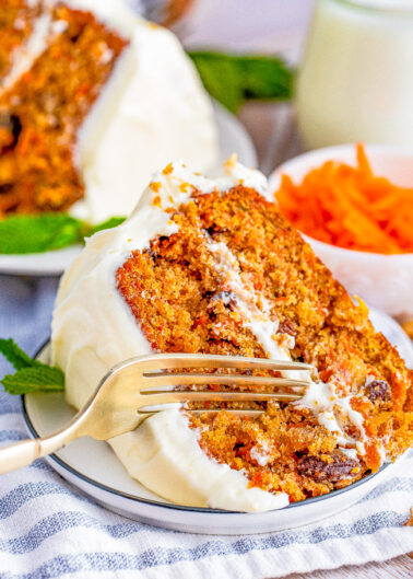 A slice of carrot cake with cream cheese frosting on a white plate, with a fork taking a bite out of it, accompanied by whole cake, shredded carrots, and a glass of milk in the background.