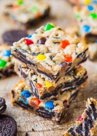 Colorful candy-studded cookie bars stacked on a wooden surface with chocolate sandwich cookies nearby.