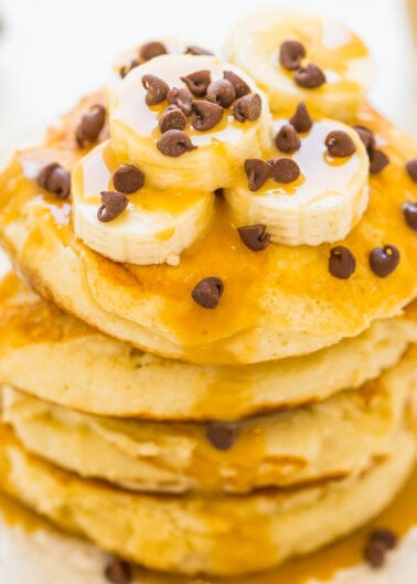 A stack of fluffy pancakes topped with banana slices, chocolate chips, and caramel drizzle.