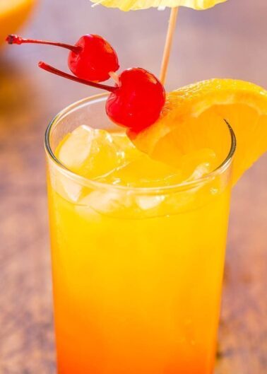 A glass of orange cocktail garnished with a slice of orange and cherries on a toothpick.