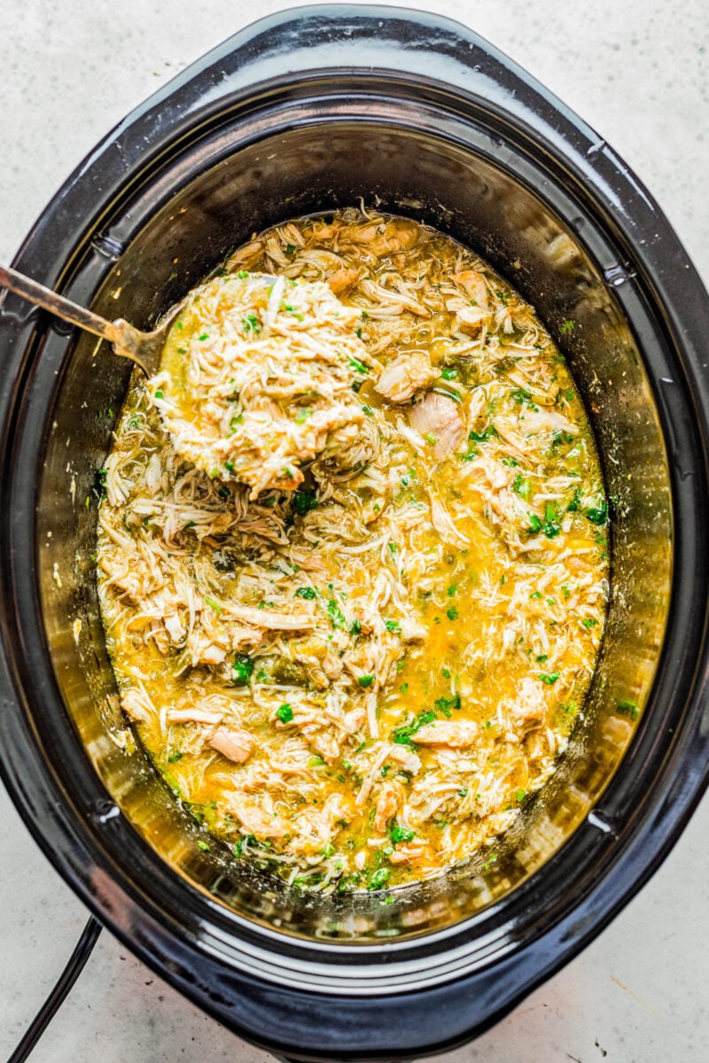 A spoon lifting a scoop of slow cooker green chile chicken from a slow cooker.