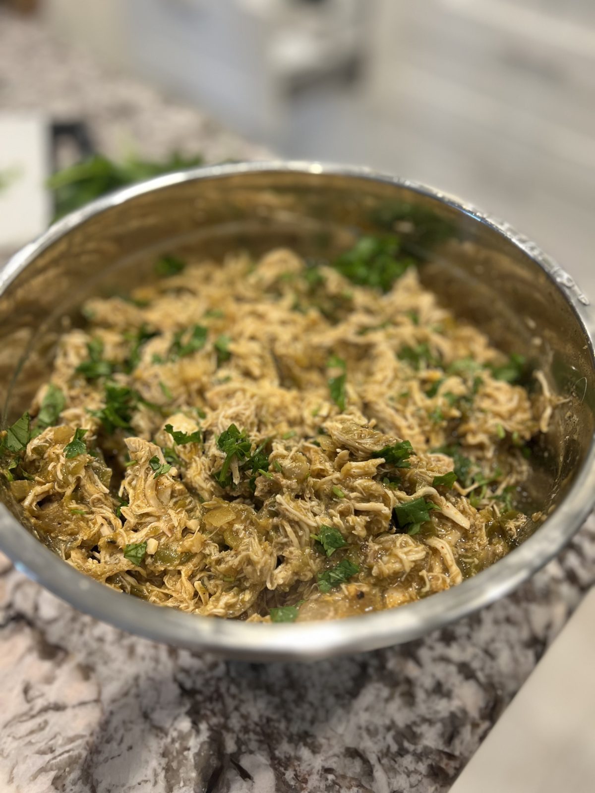 A bowl of shredded chicken mixed with chopped herbs, sitting on a marbled countertop.