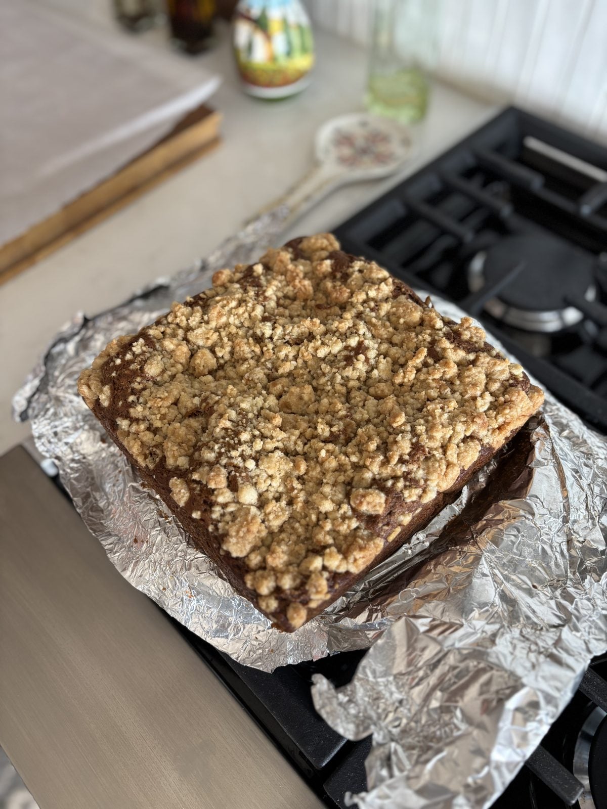 A square crumb cake on aluminum foil rests on a stovetop, with a visible crumb topping and kitchen items in the background.