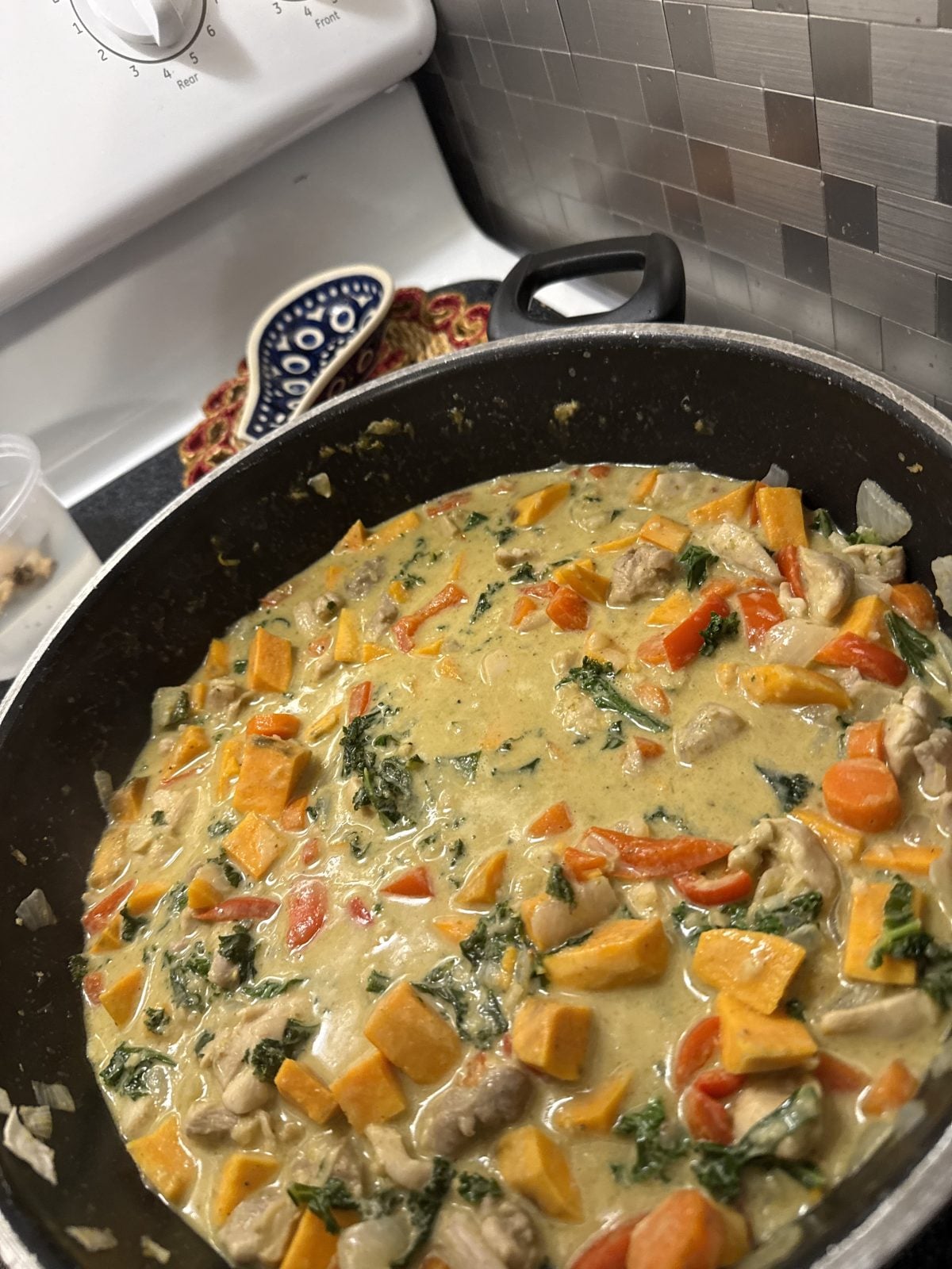A pan of stew with chunks of sweet potato, carrots, leafy greens, and pieces of meat in a creamy sauce on a stovetop.