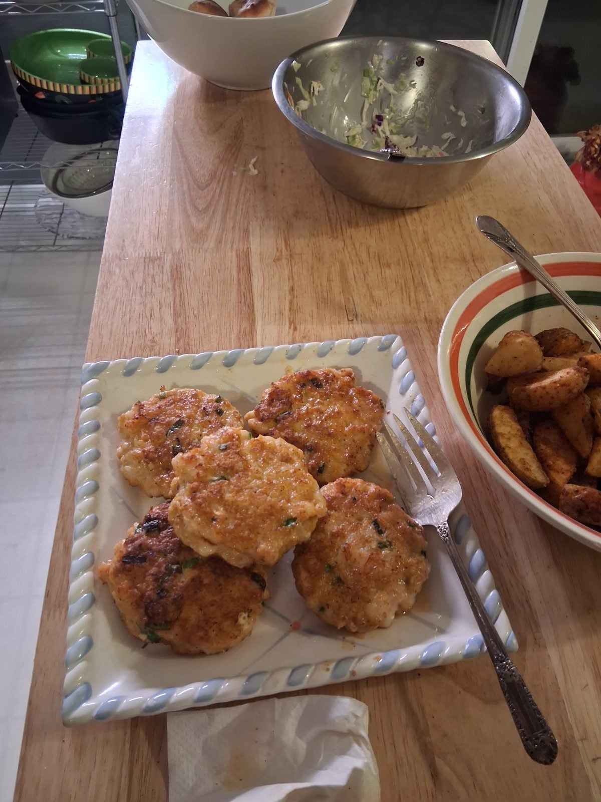 Five golden-brown patties are arranged on a square plate next to a fork. Bowls of salad and potato wedges are nearby on a wooden table.