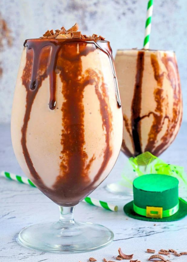Chocolate milkshake with syrup drizzle in a tall glass, next to a miniature green hat with a stripe and buckle detail.