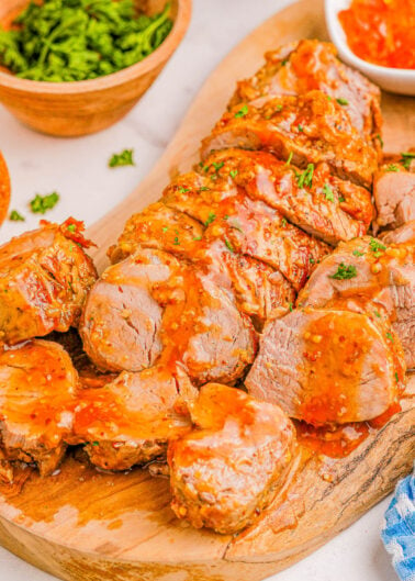 Sliced roasted pork tenderloin with glaze on a wooden cutting board, garnished with parsley.