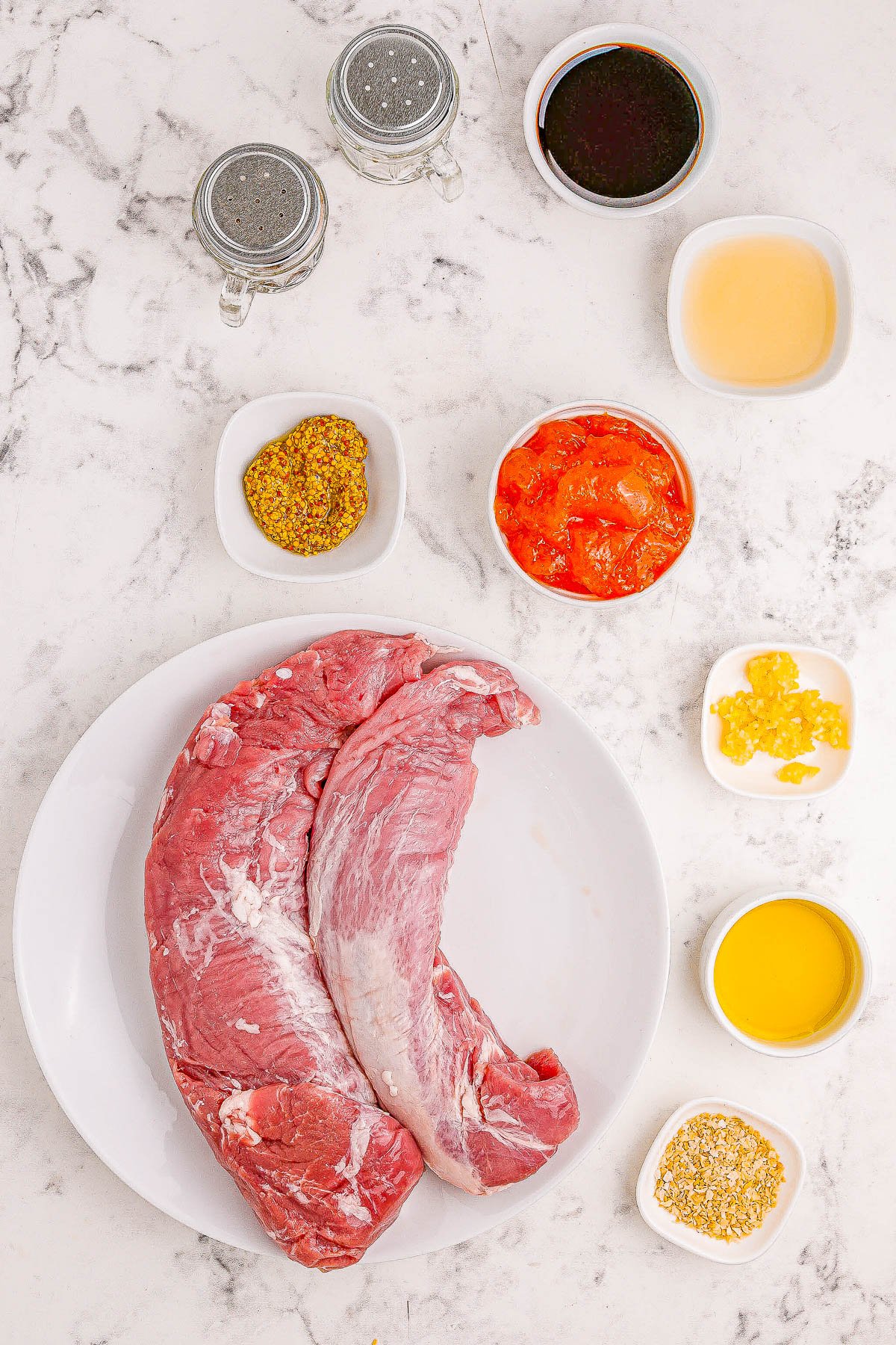 Ingredients for a beef recipe laid out on a marble countertop, including a cut of beef, various seasonings, and sauces.