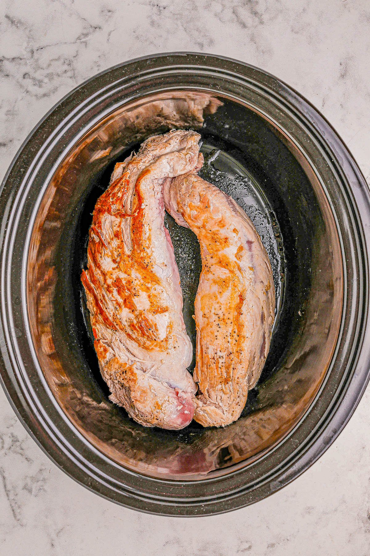 A raw, seasoned piece of meat placed inside a black slow cooker.