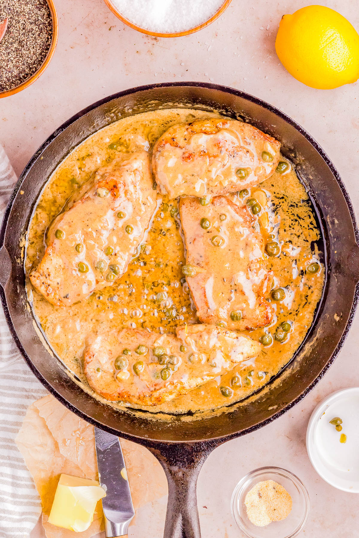 A skillet containing chicken breasts in a creamy sauce garnished with green onions, with ingredients like lemon, cheese, and spices nearby.