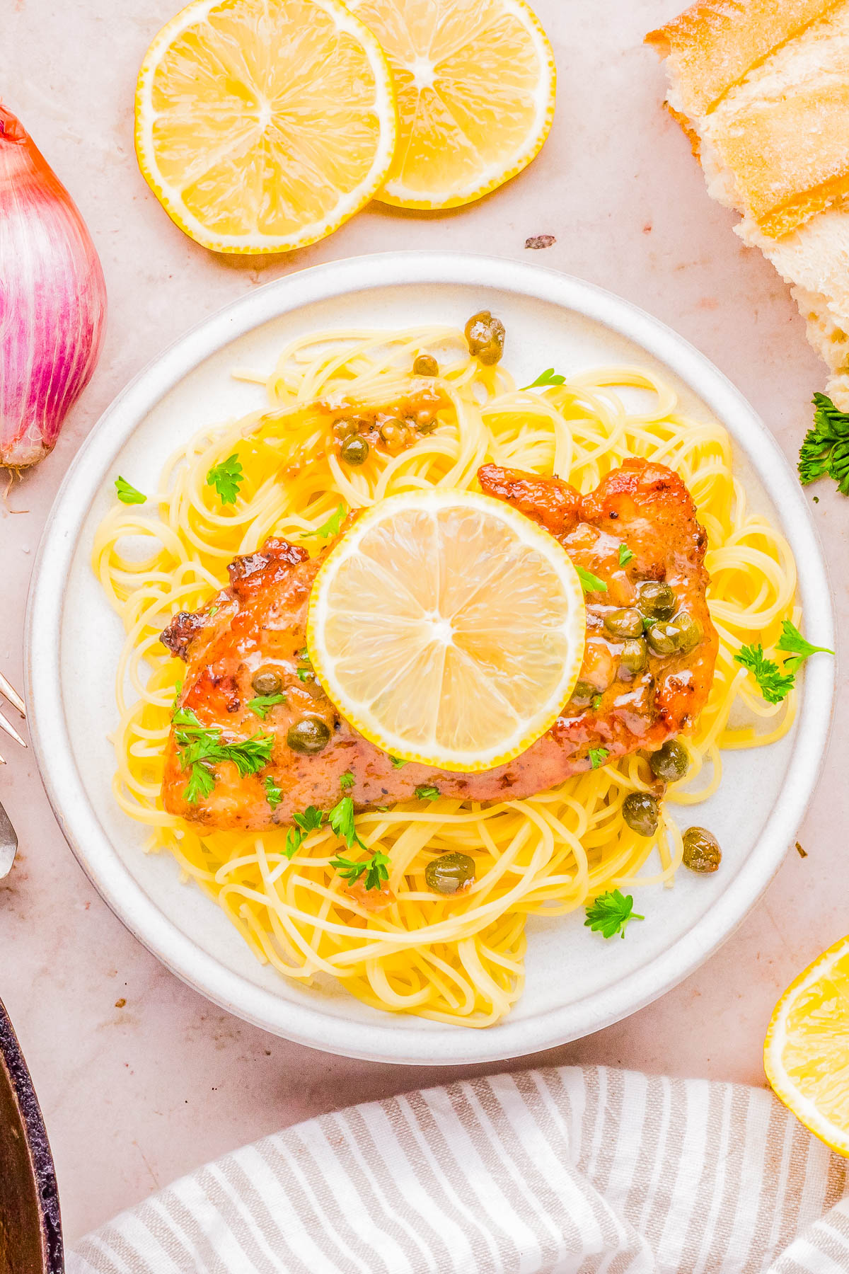 A plate of spaghetti topped with a chicken fillet, capers, and lemon slices, garnished with fresh herbs.