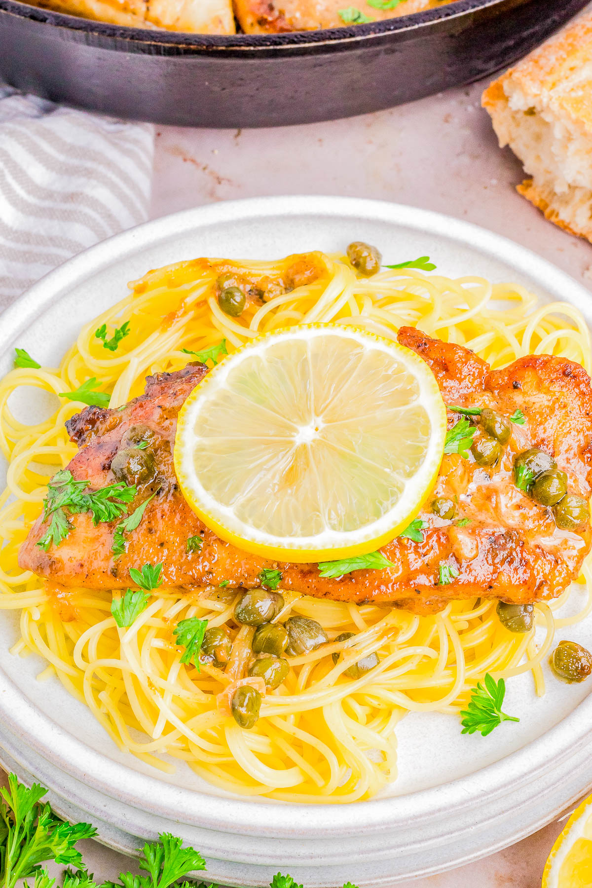 A plate of pasta topped with a grilled chicken breast, garnished with lemon slices, capers, and parsley.