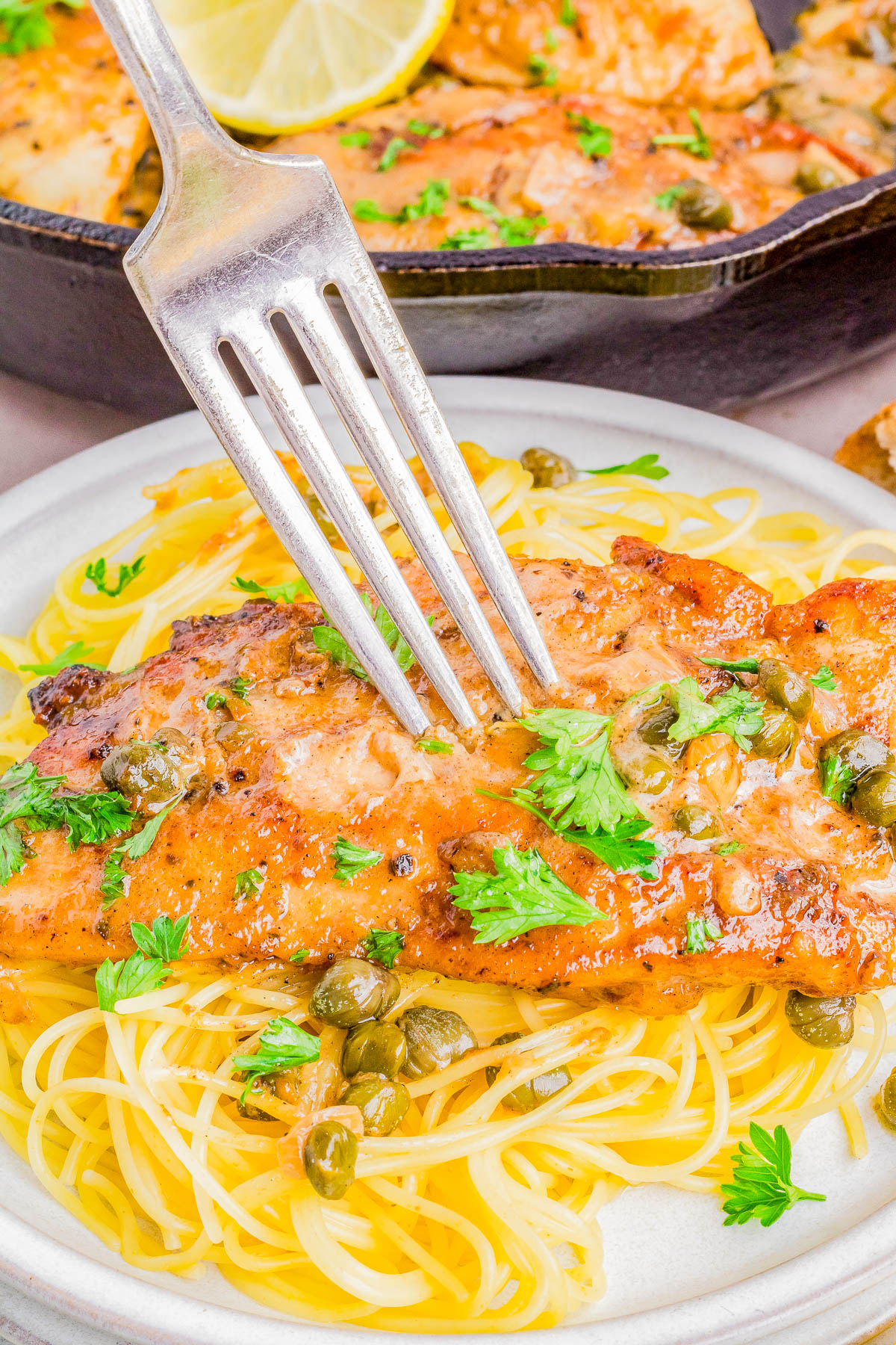 A plate of spaghetti topped with a grilled chicken breast garnished with capers and parsley, with a fork on top.