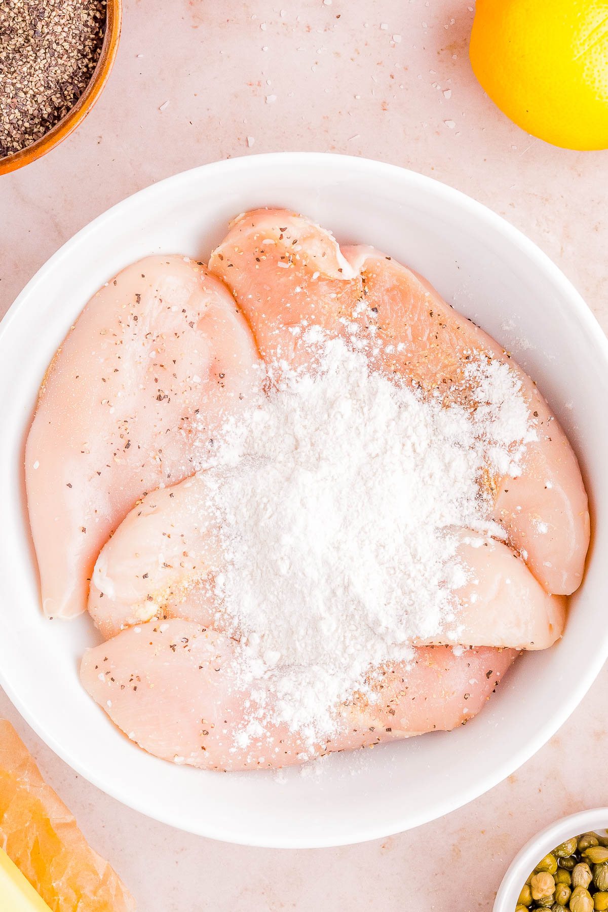 Raw chicken breasts seasoned with salt on a white plate, surrounded by cooking ingredients.