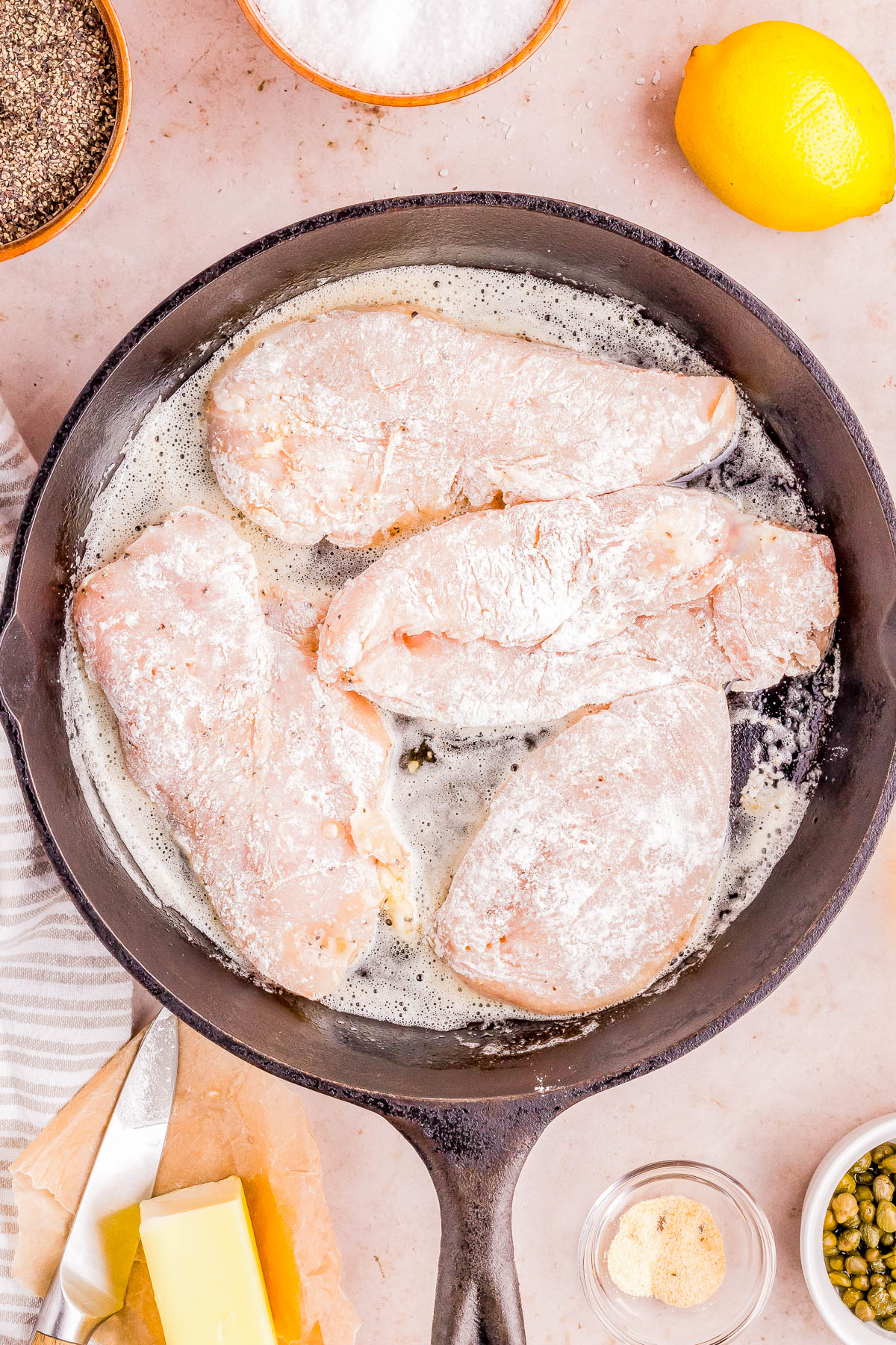 Raw chicken breasts in a seasoned flour coating being prepared for cooking in a cast-iron skillet, surrounded by ingredients like lemon, spices, and butter.