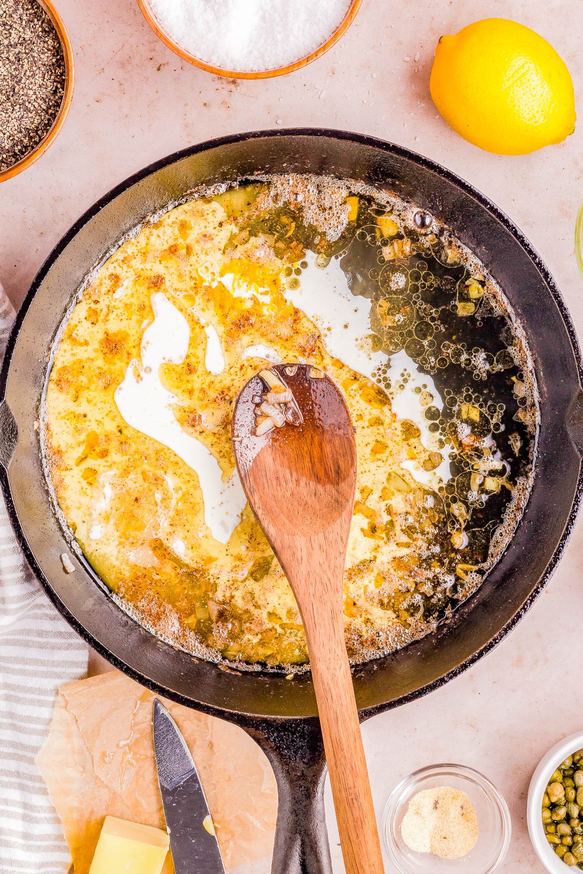 A wooden spoon resting in a skillet with bubbling butter, surrounded by ingredients like lemon, capers, and seasonings.