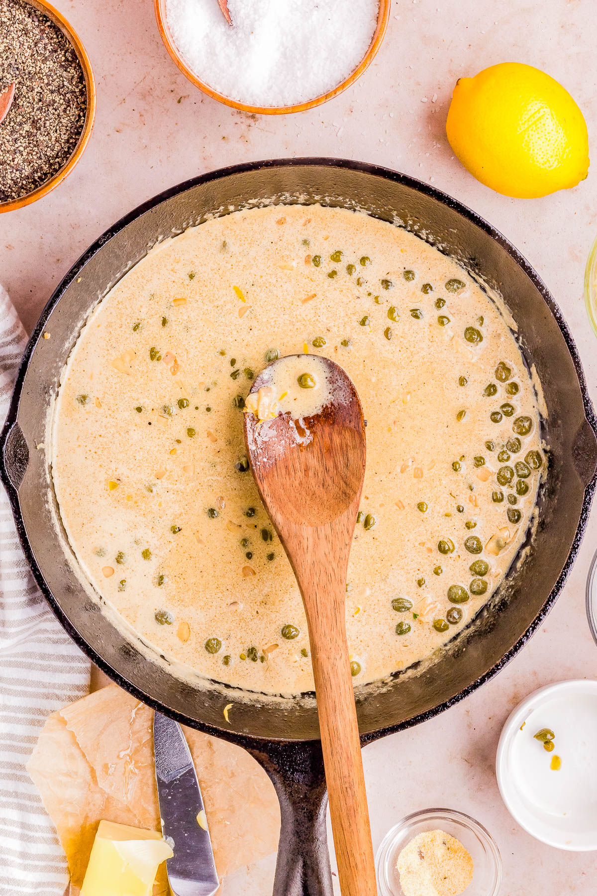 Sauce with capers being stirred in a skillet, surrounded by ingredients like lemon and spices.