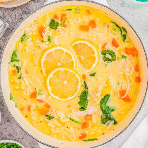 A pot of chicken noodle soup with carrot slices, noodles, and garnished with lemon slices and herbs.
