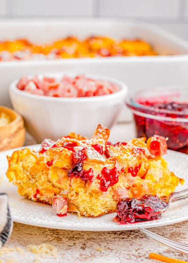 A plate of breakfast casserole garnished with diced ham and red sauce, with additional servings and condiments in the background.