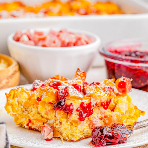 A plate of breakfast casserole garnished with diced ham and red sauce, with additional servings and condiments in the background.