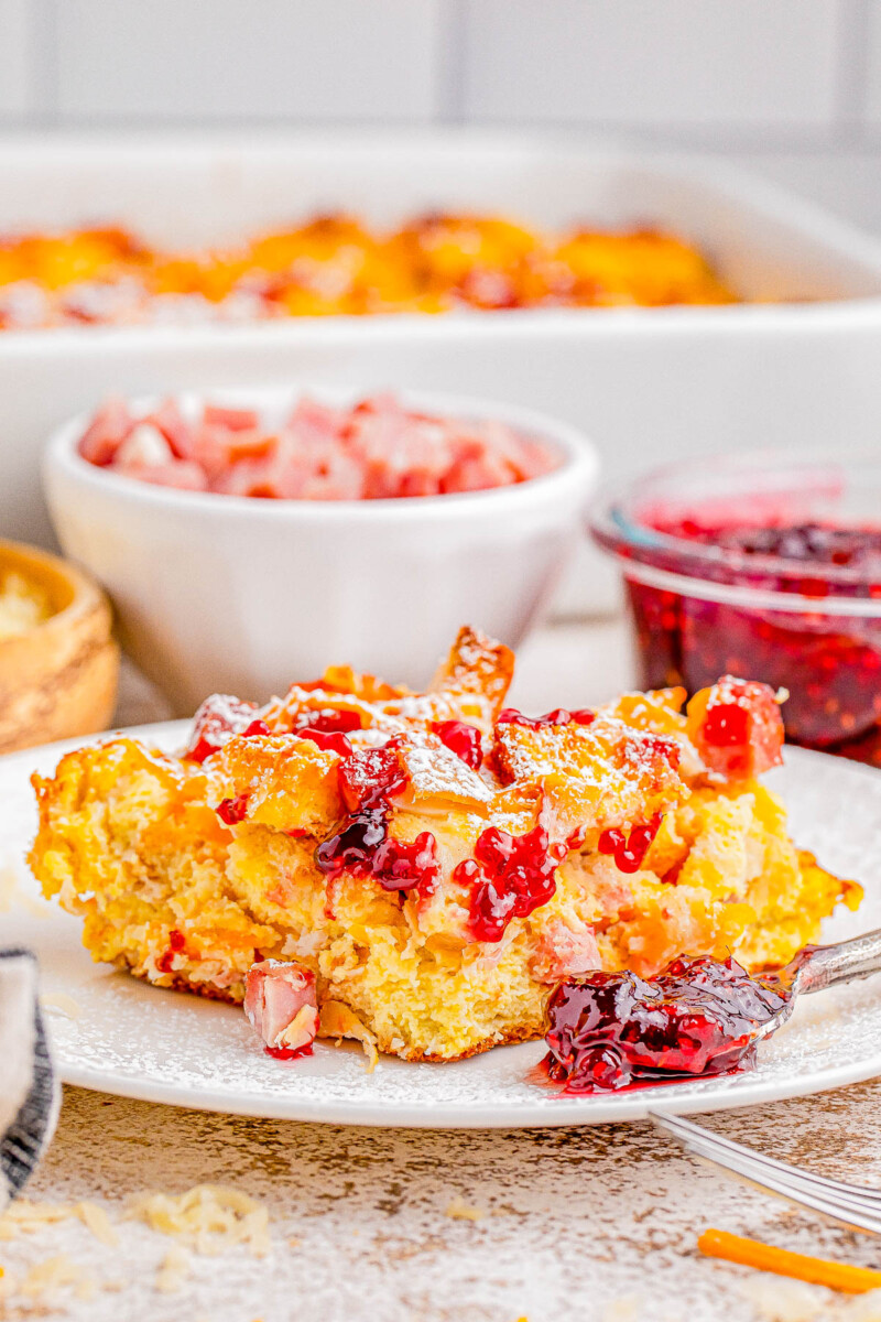 A plate of breakfast casserole garnished with diced ham and red sauce, with additional servings and condiments in the background.