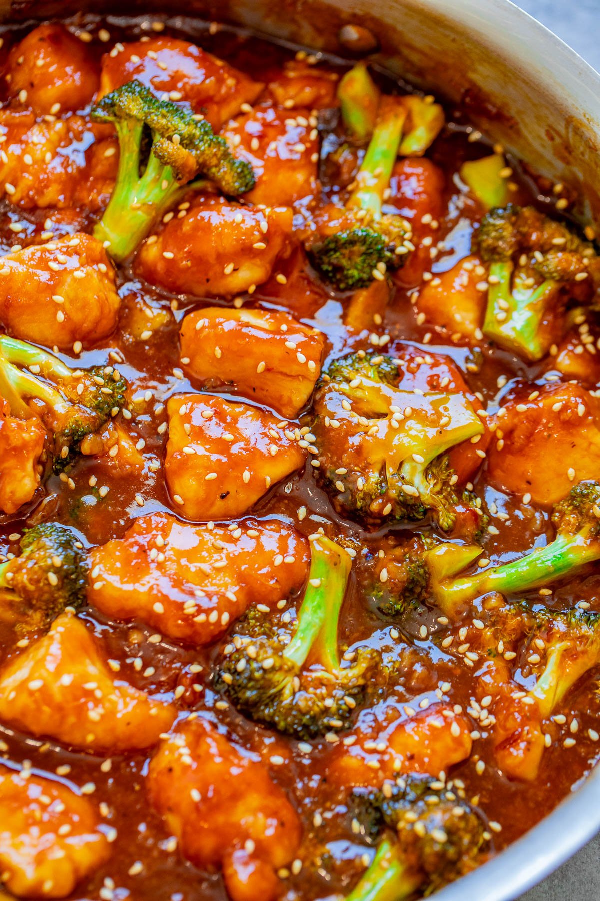 A close-up view of a dish featuring broccoli and chunks of chicken coated in a thick, glossy sauce, garnished with sesame seeds.
