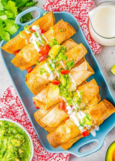 A tray of crispy taquitos topped with fresh guacamole, diced tomatoes, and sour cream, accompanied by a side of guacamole and lime wedges.