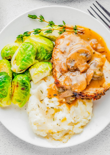 A plate of food containing sliced roasted pork with gravy, mashed potatoes, and brussels sprouts, garnished with herbs.