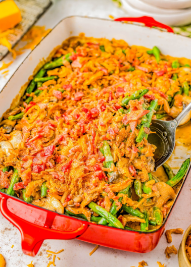 A colorful baked casserole with green beans, tomatoes, and cheese in a red dish, served with a spoon on a kitchen counter.